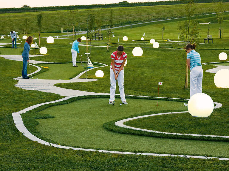 Auf einem Mini Golf Platz stehen bei einigen Bahnen Menschen und versuchen die Bälle in die Löcher zum bekommen