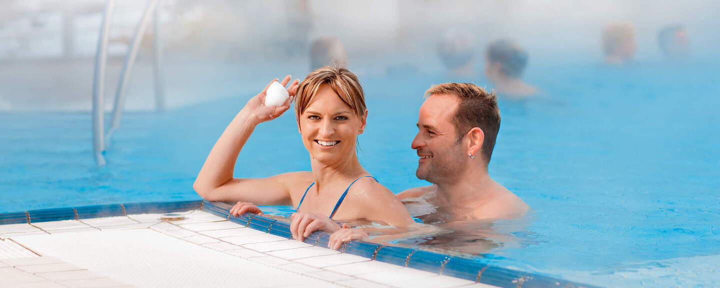 Ein Paar steht in einem Pool und die Frau wirft einen Schneeball Richtung Kamera